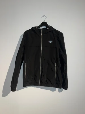 Svart vindjacka från Prada - Svart vind/regnjacka från Prada med huva och silverfärgade dragkedjor. Jackan har ett ikoniskt Prada-märke på bröstet, två fickor med dragkedja och elastiska muddar vid ärmslut och nederkant. Perfekt för dig som gillar stilren design och exklusiva detaljer.