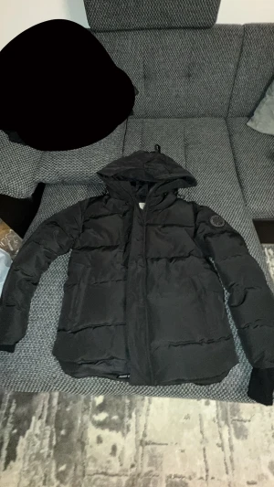 Svart pufferjacka med huva - Säljer en svart pufferjacka med huva. Jackan har quiltad design, dragkedja framtill och ribbade muddar vid ärmslut. Perfekt för kalla dagar och har en clean, modern look som funkar till det mesta. PRIS KAN DISKUTERAS!