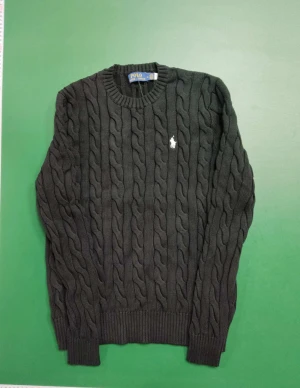 Svart stickad tröja Polo Ralph Lauren - Svart stickad tröja från Polo Ralph Lauren i storlek M. Tröjan har ett klassiskt kabelstickat mönster, rund hals och långärmade ribbade muddar. På bröstet finns den ikoniska vita Polo-loggan broderad. Perfekt för dig som gillar stilren och tidlös design.