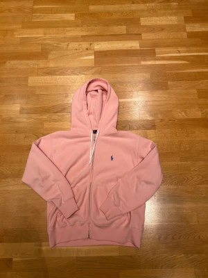 Cool hoodie från Polo Ralph Lauren - Säljer en ljusrosa hoodie från Polo Ralph Lauren med dragkedja och vit snörning vid huvan. Tröjan har ett blått broderat logomärke på bröstet, ribbade muddar och är tillverkad i mjuk bomull. Perfekt skick och köpt för 2200