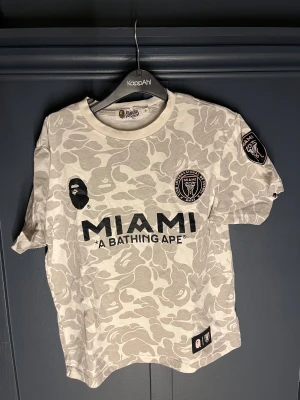 BAPE x Miami FC camo t-shirt - Säljer en limited edition BAPE x Miami FC t-shirt i ljusgrått och vitt camouflagemönster. Stor tryckt MIAMI-logga och BAPE 93 på ryggen, samt patch på ärmen. Klassisk crewneck och korta ärmar. Perfekt för dig som gillar streetwear och fotboll. Storlek medium men passar bara S