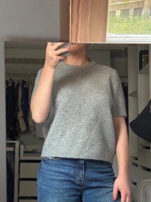 Grå stickad T-shirt - Säljer en enkel och snygg grå stickad tröja från Zara.  Tröjan har korta ärmar, rund hals och ribbade kanter vid hals, ärmslut och nederkant. Perfekt för lager-på-lager och chill stil. Materialet är mjukt och ger en skön känsla. Nopprig och lite smutsig men går att ta bort med nopperkamm och tvättas innan jag skickar!! Strl L men skulle säga att den passar S🩷