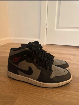 Nike Air Jordan 1 svart/grå sneakers - Säljer ett par Nike Air Jordan 1 sneakers i svart och grått med klassisk high-top siluett. Skorna har svart snörning, perforerad tå och ikonisk Swoosh på sidan. Materialet är skinn och syntet, med vit sula och röd logga på ankeln. Perfekt för dig som gillar streetwear.