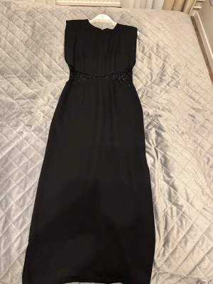 Svart långklänning med spets från Zara - Stilren svart långklänning från Zara med snygga spetsdetaljer i midjan. Klänningen har ärmlös design, rund halsringning och rak passform. Materialet är mjukt och faller snyggt. Perfekt för dig som vill ha en elegant look med en twist.