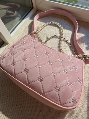 Rosa quiltad axelväska med pärldetalj - Söt rosa axelväska i quiltad sammet med små broderade rosetter. Väskan har en bred axelrem och en dekorativ kedja med pärlor som extra detalj. Perfekt storlek för det viktigaste och ger en gullig touch till din outfit.