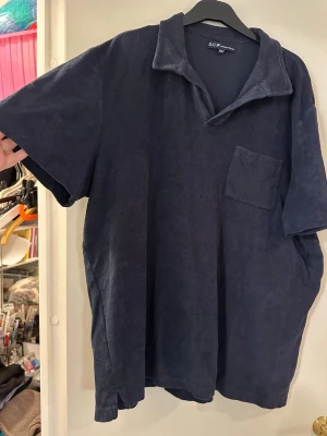 Mörkblå frottéskjorta från S.C.W XXL - Mörkblå frottetshirt från S.C.W i storlek XXL, tillverkad i mjuk frotté med 80% bomull och 20% polyester. Tröjan har korta ärmar, klassisk krage och en bröstficka. Perfekt för dig som gillar chill och oversized vibe.