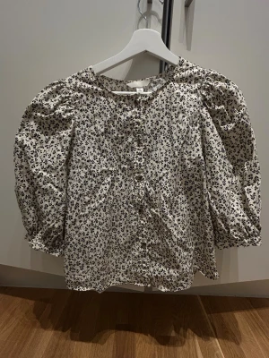 Blommig blus med puffärm från H&M - Supersöt blus från H&M i beige med svart blommönster. Blusen har puffiga trekvartsärmar, rund halsringning och knappar framtill. Tillverkad i mjuk bomull, perfekt för en trendig och avslappnad look.