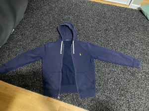 Marinblå hoodie från Polo Ralph Lauren - Snygg marinblå hoodie från Polo Ralph Lauren i storlek S. Tröjan har huva med vita snören, dragkedja framtill och två fickor. Klassisk gul broderad logga på bröstet. Perfekt för chill dagar och enkel att matcha med jeans eller joggers.