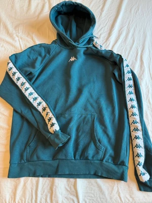Blå/grön Kappa hoodie med logoband - Snygg ”coral” hoodie från Kappa med klassiska logoband längs ärmarna och broderad logga på bröstet. Tröjan har huva med snörning och en stor magficka. Perfekt för dig som gillar sportig och avslappnad stil. Storlek M