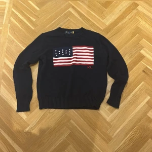 Mörkblå stickad tröja Polo Ralph Lauren - Snygg mörkblå stickad tröja från Polo Ralph Lauren med amerikansk flagga i vitt och rött på bröstet samt RL-broderi. Rund hals och ribbade muddar vid ärmslut och nederkant. Perfekt för dig som gillar klassisk preppy stil. Obs inte oriiginal