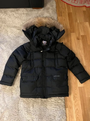 Svart Canada Goose jacka med avtagbar huva - Säljer en svart Canada Goose dunjacka som passar 13-15 år, avtagbar huva och pälsdetalj. Använd en halv vinter endast..
