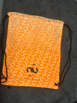 Orange Nike United ryggsäck med mönster - Snygg orange Nike United gympåse med vitt geometriskt mönster och svart logga. Väskan har svarta snören och en praktisk innerficka med dragkedja. Perfekt för att bära träningskläder eller andra grejer. Lätt och smidig design som sticker ut.