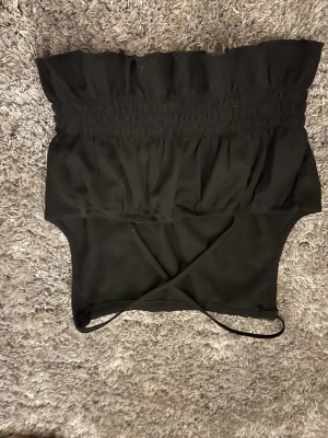 Svart croppad linnetopp med volang - Svart croppad linnetopp med tunna axelband och volangkant nedtill. Toppen har smockad midja som ger en snygg passform och en lätt rynkad struktur. Perfekt för dig som gillar stilrena och enkla plagg med en twist. Materialet känns lätt och luftigt.