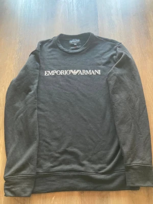 Svart sweatshirt från Emporio Armani - Svart sweatshirt från Emporio Armani med vit logga och emblem på bröstet. Klassisk rund halsringning och långa ärmar. Tillverkad i mjuk bomullsmix som känns skön mot huden. Perfekt för en clean och stilren look.