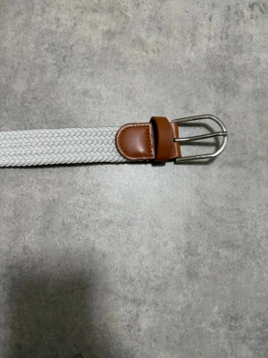 Grish bälte - Snyggt vitt flätat bälte med detaljer i brunt skinn och silverfärgat spänne. Bältet har en stilren look med klassisk form och passar perfekt till jeans eller chinos. Flätningen ger en cool struktur och gör det enkelt att justera längden.