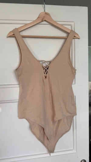 Beige bodysuit med snörning - Säljer en beige bodysuit med snygg snörning framtill. Modellen är ärmlös med breda axelband och har en rundad halsringning. Bodyn är tillverkad i mjuk bomull och har praktisk knäppning i grenen. Perfekt att styla med jeans eller kjol.