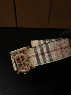 Burberry rutigt skärp med B-spänne - Säljer ett snyggt Burberry bälte med klassiskt rutigt mönster i beige, svart och rött. Bältet har ett stilrent metallspänne med Burberrys ikoniska B-logga i guldton. Tillverkat i skinn med struktur, perfekt för att lyfta din outfit.