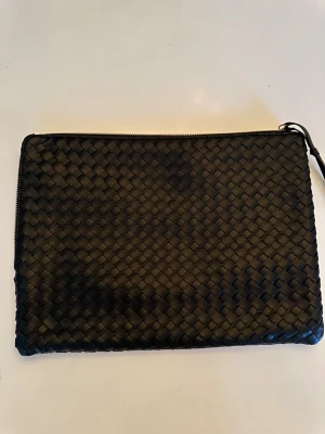 Svart flätad datorfodral i skinn - Stilren svart datorfodral med flätad design och silvrig dragkedja längs ovansidan. 