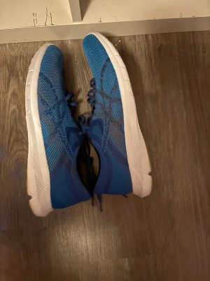 Blå Asics sneakers med mesh - Säljer ett par blå Asics sneakers med ventilerande mesh-ovandel och vita sulor med gröna detaljer undertill. Skorna har snörning och en rund tå, perfekt för dig som gillar sportig stil och vill ha bekväma skor till träning eller vardags. Snyggt Asics-tryck på innersulan.