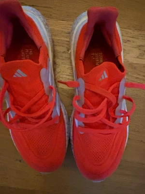 Adidas Ultraboost röda sneakers - Säljer ett par Adidas Ultraboost sneakers i starkt röd färg med vita detaljer och klassiska tre ränder på sidan. Ovandelen är i ventilerande syntetmaterial och har snörning. Sulan är tjock och dämpande, perfekt för dig som gillar att springa eller gå mycket.
