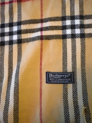 Rutig beige halsduk från Burberrys - Klassisk halsduk från Burberrys i beige med rutigt mönster i svart, vitt och rött. Gjord av 100% lammull och har fransar i ändarna. Perfekt för att ge din outfit en tidlös och lyxig touch. Märke och materialetikett synlig på ena sidan.