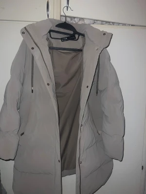 Beige jacka från Zara - Säljer en beige jacka från Zara med huva och dragkedja framtill. Jackan har långa ärmar, snörning vid huvan och praktiska fickor på sidorna. Perfekt för kalla dagar och har en clean, modern look.