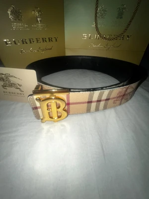 Burberry bälte - Säljer ett Burberry bälte med klassiskt randigt mönster i beige, brun och röd. Bältet har ett stort guldigt 'B'-spänne och är tillverkat i skinn med textil på utsidan. Perfekt accessoar för dig som vill sticka ut med ett lyxigt bälte! Bra skick på bältet. Ny pris ca:4000 kr. vitto finns😁
