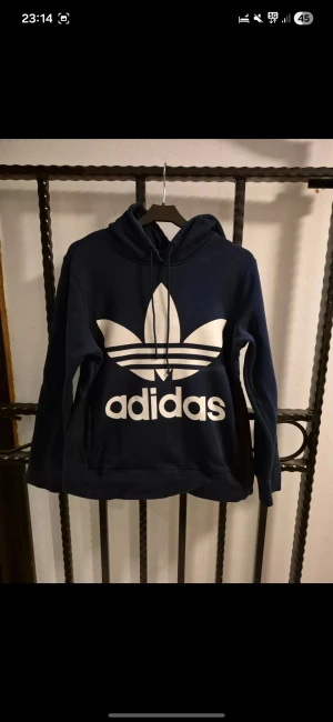 Marinblå Adidas hoodie med logga - Snygg marinblå hoodie från Adidas med stor vit trefoil-logga och text på bröstet. Tröjan har huva med snörning och långa ärmar. Mjuk insida i bomullsmix som gör den skön att bära. Perfekt för dig som gillar streetwear och sportig stil.