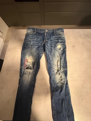 Dsquared2 blå jeans med slitningar - Snygga blå jeans från Dsquared2 med coola slitningar, färgstänk och patchdetaljer på benen. Klassisk femficksmodell med läderpatch bak och röd Dsquared2-tag vid fickan. Jeansen har en rak passform och är tillverkade i robust denim.