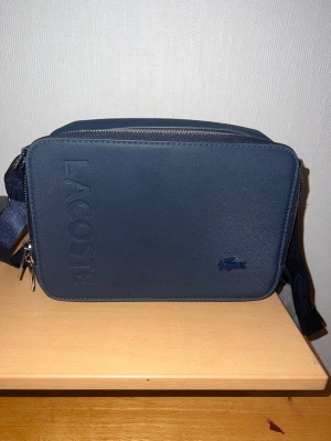 Mörkblå axelväska från Lacoste - Snygg mörkblå axelväska från Lacoste med stilren design och präglad logga på framsidan. Väskan har en praktisk dragkedja och är tillverkad i slätt skinnmaterial. Perfekt storlek för att bära med sig det viktigaste och passar till en modern streetstil.