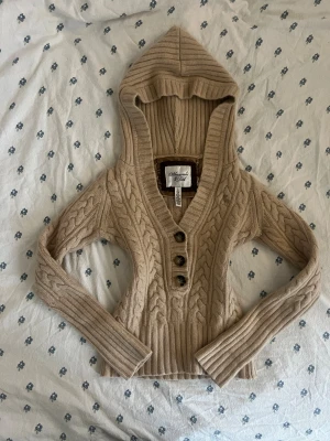 Vintage beige stickad hoodie Abercrombie - Vintage beige stickad hoodie från Abercrombie & Fitch med flätmönster, djup v-ringning och tre snygga knappar framtill. Tillverkad i ull, rayon & kaninhår. Vintage storlek L passar mer som xs. Lite noppriga & märkesetiketten börjar lossna lite men inget som inte kan lagas eller påverkar slitaget. Säljer pga för liten. Ha inga bilder med den på. Hade också bytt mot en lik. Ungefär mått (platta): Axel till axel: 36cm Axel till botten: 47cm Armhåla till armhåla: 37cm Yttre arm: 62cm Inre arm: 47cm