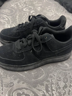 Nike Air Force 1 Low Svart Mocka - Säljer ett par svarta Nike Air Force 1 Low i mocka med klassisk snörning och rund tå. Skorna har en platt sula och svart Nike Air-logga på hälen. Stilrena och tidlösa sneakers med mjukt textilfoder och snygga detaljer i svart.