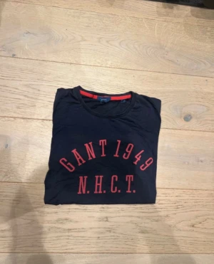 Svart GANT t-shirt med röd text - Cool svart t-shirt från GANT med trycket 'GANT 1949 N.H.C.T.' i rött framtill. Rund hals med röd kant på insidan ger en snygg detalj. Passar dig som gillar klassisk streetstil och tydliga loggor. Mjuk och skön bomullskvalitet.