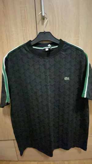 Svart mönstrad t-shirt Lacoste S - Svart t-shirt från Lacoste i storlek S med geometriskt mönster över hela plagget. Klassisk rund hals och korta ärmar med gröna och vita ränder längs axlarna. Ikoniska Lacoste-loggan i grönt broderad på bröstet. Perfekt för en sportig och stilren look.