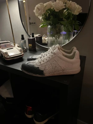 Maison Margiela sneakers svart/vit - Maison Margiela sneakers i vitt skinn med svart tå och coolt stänkmönster över hela skon. Klassisk låg modell med snörning och rund tå. Snygg och unik design som sticker ut, perfekt för dig som gillar statement sneakers.