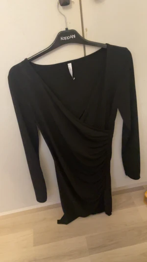 Svart omlottklänning - Svart långärmad omlottklänning med snygg drapering i midjan och v-ringad hals. Klänningen har en figurnära passform och är perfekt för dig som gillar stilrena och eleganta plagg.