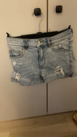 Ljusblå denimshorts med slitningar - Säljer ett par ljusblå denimshorts med coola slitningar och uppvikta ben. Klassisk femficksmodell med knapp och dragkedja framtill. Perfekta för dig som gillar en avslappnad och trendig stil på sommaren.