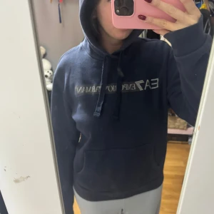 Mörkblå hoodie från Emporio Armani - Säljer en mörkblå hoodie från Emporio Armani EA7 med logga i silver på bröstet. Tröjan har huva med snörning, stor magficka och långa ärmar. Materialet är mjuk bomull och polyester, perfekt för chill dagar. Snygg och enkel design som funkar till jeans eller mjukis.