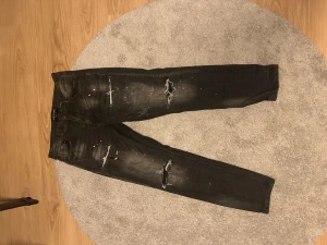 Svarta jeans Supply & Demand, 32W - Svarta jeans från Supply & Demand med slitna detaljer och färgstänk. Jeansen har smal passform, klassisk femficksdesign och är tillverkade i ett stretchigt bomullsmaterial. Använda endast en gång