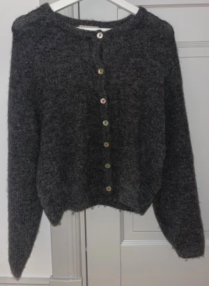 Mörkgrå stickad kofta med knappar - Mysig mörkgrå stickad kofta från Ginatricot med rund hals och snygga knappar framtill. Koftan har långärmade ärmar och en lite loose passform som gör den extra skön. Perfekt att slänga på sig när det är kyligt ute.