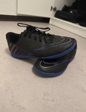 Svarta Nike fotbollsskor med blå sula - Säljer ett par svarta Nike fotbollsskor med blå sula och snygga silverdetaljer. Skorna har snörning och en låg profil, med ett stilrent Nike-tryck på sidan. Perfekt för inomhusspel och har en platt sula för bra grepp på planen.