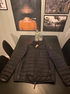 Svart dunjacka från Parajumpers - Säljer en svart dunjacka från Parajumpers med klassisk patch på ärmen och gul detalj vid halsen. Jackan har quiltad design, hög krage och dragkedjor på fickorna. Perfekt för dig som vill ha en lätt men varm jacka med snygg passform. Har dock en liten laglapp vid patchen som knappt syns Storlek M