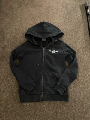 Svart hoodie Peak Performance 170 - Svart hoodie från Peak Performance med vit logga på bröstet. Tröjan har dragkedja, huva och två fickor framtill. Mjuk insida och ribbade muddar vid ärmslut och nederkant. Perfekt för chill eller träning.