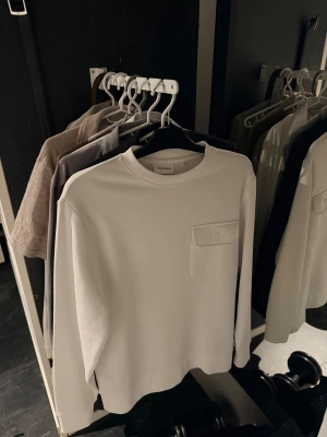 Vit långärmad tröja från Lindbergh ( HELT NY ) - Vit långärmad tröja från Lindbergh med rund hals och en snygg bröstficka med knapp. Enkel och clean design som funkar till många olika stilar. Tröjan är helt ny och aldrig använd!