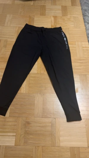 Svart sweatshirt från BOSS - Svart sweatshirt från BOSS med silverfärgad rand och logga över bröstet. Tröjan har rund hals och långa ärmar, tillverkad i mjuk bomull som känns skön mot huden. Perfekt för dig som gillar stilren och enkel design med en touch av street.