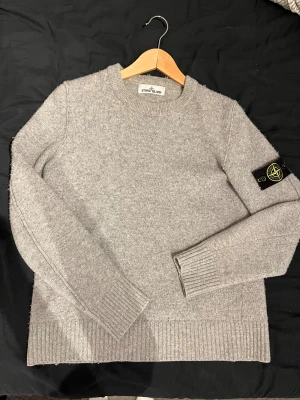 Grå stickad Stone Island tröja - Snygg grå stickad tröja från Stone Island med klassisk rund hals och ribbade muddar. Tröjan har lång ärm och den ikoniska Stone Island patchen på vänster ärm. Perfekt för dig som gillar stilrena och exklusiva plagg. Plagget passar mera en Medium/small men är en large