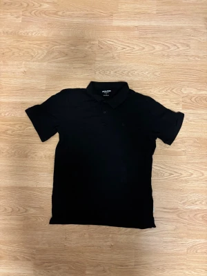 Svart pikétröja från Jack & Jones - Klassisk svart pikétröja från Jack & Jones med korta ärmar och krage med två knappar. Tröjan är gjord i mjuk bomull och har en stilren look som funkar till många olika outfits. Perfekt för dig som gillar enkel och clean stil.