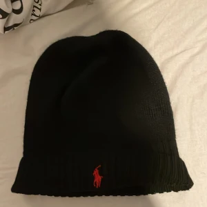 Svart mössa från Polo Ralph Lauren - Svart ribbstickad mössa från Polo Ralph Lauren med klassisk röd broderad logga framtill. Mjuk och skön passform, perfekt för kalla dagar. Stilren design som funkar till alla jackor och outfits.