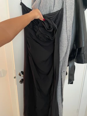 Svart långklänning från MNG Capsule - Elegant svart långklänning från MNG Capsule med en unik stor blomdetalj framtill. Klänningen har en smickrande drapering och är tillverkad i ett mjukt, följsamt material som ger en lyxig känsla. Perfekt för dig som vill sticka ut med en stilren och trendig look.