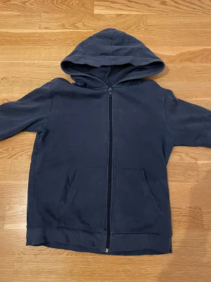 Mörkblå hoodie med dragkedja - En enkel och stilren mörkblå hoodie med huva och hel dragkedja framtill. Tröjan har två praktiska fickor och är tillverkad i mjukt fleece-material. Perfekt för chill dagar eller när du vill ha något bekvämt och varmt på dig.